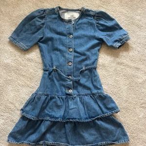Jeansklänning från Les Coyotes - Supersnygg jeansklänning från Les Coyotes med kort ärm, knappar framtill och volangkjol. Klänningen har markerad midja med bälte och puffärm. Perfekt nu i sommar