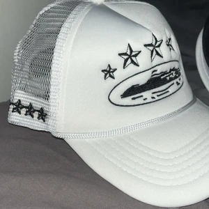 Corteiz keps  - Snygg vit truckerkeps med svart broderade stjärnor och motiv framtill. Kepsen har mesh på sidorna och bak för extra ventilation och en klassisk böjd skärm. Perfekt accessoar för en cool streetstyle.