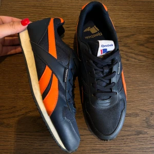 Reebok sneakers i svart och orange - Snygga Reebok sneakers i svart med orange detaljer och vit sula. Klassisk retrostil med mesh och syntetmaterial, snörning framtill och rund tå. Perfekta för dig som gillar sportig och färgstark look. STORLEK 38,5 