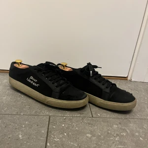 Svarta Saint Laurent sneakers - Svarta sneakers från Saint Laurent med broderad logga på sidan. Skorna har en klassisk låg profil, svarta snören och beige sula. Hör av er vid frågor eller funderingar! Kan gå ner i pris vid snabb affär 