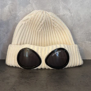 C.P. Company Vit mössa - Vit ribbstickad mössa från C.P. Company med ikoniska svarta goggle-linser framtill. Mössan är tillverkad i mjuk ull och har en bred uppvikt kant. Perfekt för dig som vill sticka ut med streetwear-vibe och cool detalj. Köptes sprillans ny på johnells i vintras. Säljes pga att den inte kommer användas.