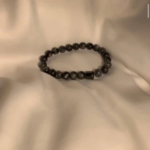 Svart pärlarmband med stenar - Stilrent armband med svarta och gråmelerade pärlor i rund form. Pärlorna är trädda på elastisk tråd och har en diskret metallisk detalj. Perfekt accessoar för en cool och enkel look.