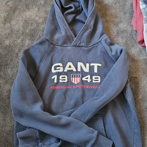 Blå hoodie från GANT med brodyr - Mörkblå hoodie från GANT med broderad logga och texten '1949 American Sportswear' på bröstet. Tröjan har huva, känguruficka och ribbade muddar. Perfekt för dig som gillar sportig och avslappnad stil.