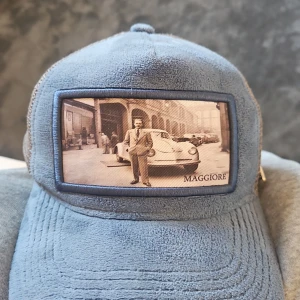 Blågrå keps från Maggiore med mesh - Snygg blågrå keps från Maggiore med vintageinspirerad patch framtill. Kepsen har mesh på sidorna och bak, samt justerbart spänne i skinnimitation. Perfekt för dig som gillar streetstyle och unika detaljer.