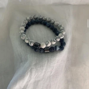 Två pärlarmband i blått och vitt - Två elastiska armband med pärlor, ett med vita pärlor med grå marmorering och ett med mörkblå pärlor med spräckligt mönster. Båda har detaljer i metallfärg. Perfekt accessoar för att lyfta din stil.