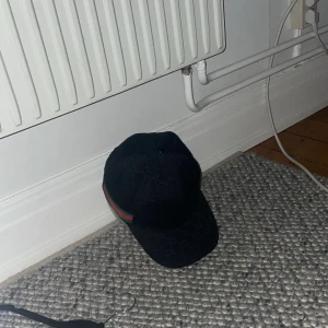 Svart keps från Gucci med logomönster - Snygg svart keps från Gucci med diskret GG-logomönster över hela kepsen och klassiska gröna och röda ränder på sidan. Justerbar rem baktill i svart skinn. Tillverkad i Italien och riktigt lyxig känsla. Perfekt accessoar för att lyfta din outfit.