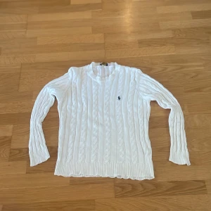 Vit kabelstickad tröja från Ralph Lauren - Vit kabelstickad tröja från Ralph Lauren med rund halsringning och broderad logga på bröstet. Tröjan har långa ärmar och ribbade muddar vid ärmslut och nederkant. Perfekt för en klassisk och stilren look.