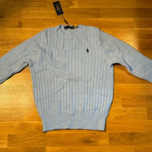 Ljusblå stickad tröja från Polo Ralph Lauren - Snygg ljusblå stickad tröja från Polo Ralph Lauren med klassiskt kabelstickat mönster och broderad logga på bröstet. Tröjan har rund halsringning och långa ärmar, perfekt för lager-på-lager-stil. Den är   M men passar XS/S