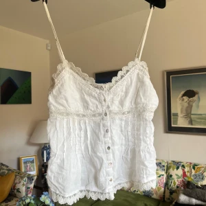 Vit linnetopp från Zara - Supersöt vit linnetopp med tunna axelband, knappar framtill och fina spetsdetaljer längs kanterna. Toppen har en romantisk känsla med broderad spets och lätt rynkade partier. Perfekt för varma dagar eller lager på lager.