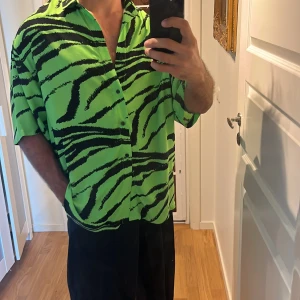 Grön zebramönstrad skjorta ASOS - Säljer en riktigt fet grön skjorta från ASOS DESIGN med svart zebramönster. Skjortan har korta ärmar, knappar framtill och en loose fit som ger en avslappnad vibe. Perfekt för dig som vill sticka ut med en unik look.