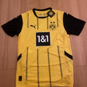 Borussia Dortmund matchtröja Puma M - Gul och svart Borussia Dortmund fotbollströja från Puma med svarta vertikala ränder, klubbmärke och sponsortryck på bröstet. Tillverkad i lätt och ventilerande material (RE:FIBRE). Klassisk rund hals och korta ärmar. Perfekt för dig som älskar BVB!