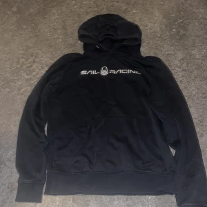 Svart Sail Racing hoodie med logga - Svart hoodie från Sail Racing i storlek M. Klassisk modell med huva, stor magficka och broderad logga på bröstet. Mjuk insida och stilren design med Sail Racing-tryck framtill. Perfekt för dig som gillar sportig och avslappnad stil.