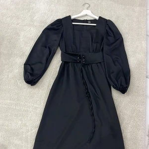 Svart långklänning  - Svart långklänning från SHEIN med puffiga långärmar, fyrkantig ringning och bred midjedetalj med snörning. Satin. Storlek S