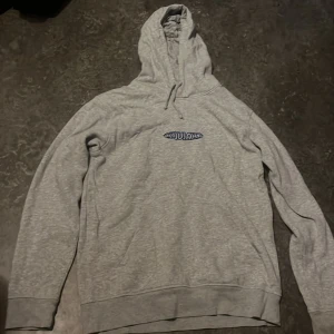 Grå hoodie från Tjuvjakt med brodyr - Säljer en grå hoodie från Tjuvjakt med blå broderad logga på bröstet. Tröjan har huva med snörning och långa ärmar. Perfekt för dig som gillar streetwear och vill ha något bekvämt och stilrent.