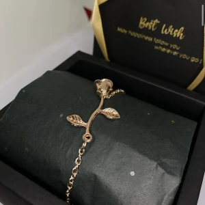 Guldigt armband med rosdetalj - Snyggt armband i guldfärg med en detalj i form av en ros och två blad. Kedjan är tunn och smidig, perfekt för att ge en romantisk touch till din stil. Kommer i en elegant presentask, redo att ges bort eller behålla själv. Ej använd utan kommer från min small business. Finns även i Silver. Skriv om ni vill köpa med matchade halsband. 