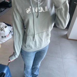 Ljusgrå/blå Levi's hoodie med tryck - Säljer en ljusgrå hoodie från Levi's med vit logga. Perfekt för dig som gillar en clean och avslappnad stil. Materialet är mjuk bomull som känns skönt mot huden. 
