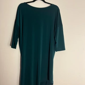 Mörkgrön långklänning med trekvartsärm - Enkel och stilren mörkgrön långklänning med trekvartsärm och rund halsringning. Klänningen har en rak och loose passform med något längre bakstycke. Materialet känns mjukt och följsamt, perfekt för dig som gillar minimalistisk stil. Finns en liten fläck.