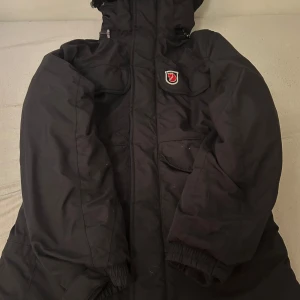 Svart vinterjacka från Fjällräven - Svart vinterjacka från Fjällräven med klassisk röd räv-logga på bröstet. Jackan har flera stora fickor framtill, hög krage och huva. Tillverkad i slitstark polyester och har långa ärmar med resår vid handlederna. Perfekt för kalla dagar och outdoor vibes.