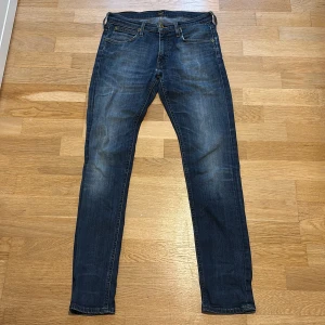 Lee Luke jeans W30 L32 blå - Säljer ett par Lee Luke jeans i klassisk blå tvätt med snygga slitningar. Modellen har smal passform och raka ben, med Lee-logga på bakfickan och läderpatch i midjan. Perfekta för dig som gillar en modern och stilren look.