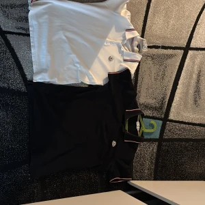 Moncler pikétröjor svart & vit M - Två stilrena Moncler pikétröjor i svart och vitt. Båda har klassisk krage med röd och blå rand, broderad Moncler-logga på bröstet och knäppning framtill. Perfekta för dig som gillar exklusiva och sportiga vibes. Materialet är mjuk bomull som andas.