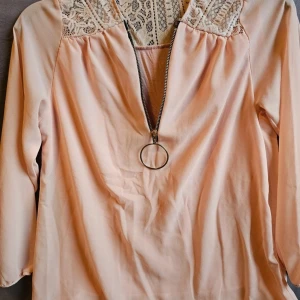 Ljusrosa/aprikos blus med fina detaljer  - Supersöt ljusrosa/aprikos blus från MYHOL S i storlek L. Blusen har spetsdetaljer upptill, en stor rund dragkedja framtill och trekvartsärmar. Perfekt för dig som gillar romantiska och trendiga detaljer.