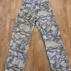 Zara jeans med camouflage - Jeans från Zara med ett iögonfallande kamouflagemönster i jordnära toner av beige, grått och svart som påminner om skogsbark och naturens strukturer. Byxorna har en avslappnad, rak passform och är tillverkade i ett tåligt denimliknande material som passar både till streetwear och utomhusaktiviteter. De är designade med flera praktiska fickor, både framtill och baktill, inklusive en klassisk verktygsficka på sidan. 