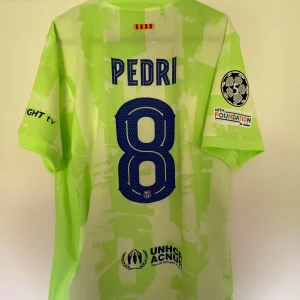 FC Barcelona Pedri bortatröja Nike - Säljer en FC Barcelona bortatröja med Pedri 8 på ryggen. Tröjan är ljusgrön med abstrakt mönster, officiella tryck och Champions League-patch på ärmen. Nike-logga, Spotify och UNHCR-tryck framtill. Tillverkad i lätt och ventilerande material.