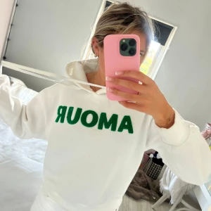 Vit hoodie med grön AMOUR-text - Säljer en vit hoodie med huva och snörning. På bröstet står det AMOUR i stora gröna bokstäver med en fluffig textur. Hoodien har en klassisk passform och är perfekt för en chill och avslappnad stil. Den har en lite fläck på högra sidan men syns inte tydligt!