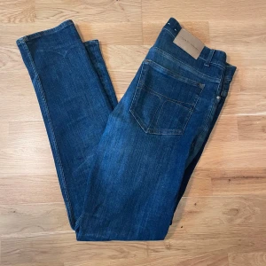 Mörkblå jeans Evolve 34/32 - Snygga mörkblå jeans från Tiger of Sweden i modellen Evolve. Klassisk femficksdesign, raka ben och normal passform. Jeansen är tillverkade i ett slitstarkt bomullstyg med lätt tvättad look och subtila kontrastsömmar. Hör av er för mått eller andra frågor om jeansen!