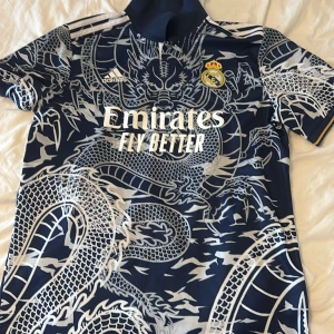 Real Madrid drakmönstrad fotbollströja - Snygg Real Madrid fotbollströja från Adidas med stort drakmönster i vitt på marinblå botten. Tröjan har klubbmärke och Adidas-logga på bröstet samt Emirates-sponsring. Kortärmad modell med klassisk krage och sportig känsla. Perfekt för dig som vill sticka ut på planen.
