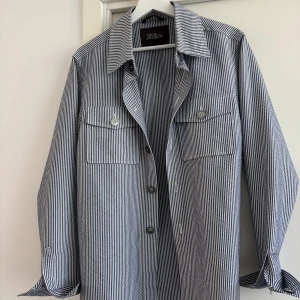 Randig overshirt från Oscar Jacobson - Snygg randig overshirt från Oscar Jacobson i blått och vitt. Jackan har klassisk skjortkrage, två bröstfickor med knappar och långa ärmar med manschett. Perfekt lager-på-lager-plagg för vår och sommar. Tillverkad i bomull för en lätt och luftig känsla. Nypris 3599