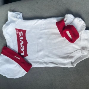 Vit Levi's body med rött tryck - Supersöt vit body från Levi's med klassiskt rött logotryck framtill. Setet inkluderar matchande vit mössa och vita strumpor med röda muddar, båda med Levi's-logga. Perfekt för en liten fashionista som vill sticka ut med ikonisk streetstyle. Strl 62 