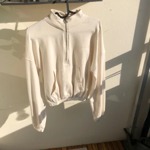 Beige half zip croppad hoodie  - Snygg beige croppad hoodie med half zip framtill och broderad logga på bröstet. Tröjan har lång ärm, känguruficka och elastisk midja jätte skön lite tunnare material o luftig