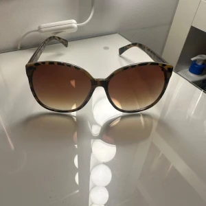 Leopardmönstrade solglasögon Jessica Simpson - Stora solglasögon från Jessica Simpson med leopardmönstrad båge i bruna och svarta toner. Glasen är bruna och tonade, vilket ger en snygg och trendig look. Perfekta för att lyfta din stil under soliga dagar.