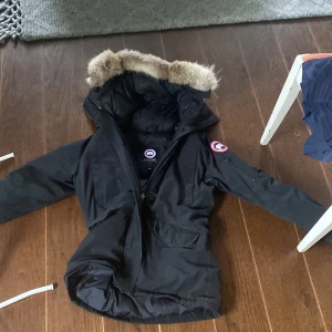 Svart Canada Goose dunjacka med päls - Säljer en svart Canada Goose dunjacka i storlek S med ikonisk patch på ärmen och fluffig päls runt huvan. Jackan har dragkedja, stora fickor och är perfekt för kalla vinterdagar. Klassisk och snygg modell som håller dig varm hela säsongen. Storleken är S men sitter mer som en xs (kan passa båda) dragkedjan på jackan är lite trög och den är mycket använd. Priset kan varieras. Hör av dig vid frågor!