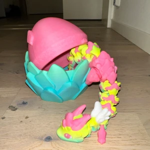 Färgglad drakleksak med ägg - En färgglad drakleksak som kommer med ett matchande ägg. Draken har en kropp i rosa, gult och grönt med vita detaljer och kan förvaras i det turkosa och rosa ägget. Perfekt för lek och samling!