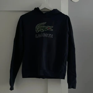 Svart hoodie från Lacoste - Säljer en svart hoodie från Lacoste med den ikoniska krokodilloggan på bröstet. Tröjan har långa ärmar och en klassisk huva. Perfekt för en avslappnad stil.