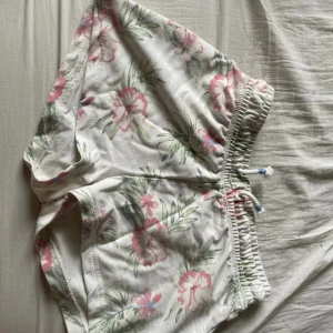 Blommiga shorts från Cubus - Säljer ett par vita shorts med blommigt mönster i rosa och grönt från Cubus. De är tillverkade i en mjuk blandning av bomull och polyester. Shortsen har en elastisk midja med knytband för extra komfort. Perfekta för en avslappnad stil!