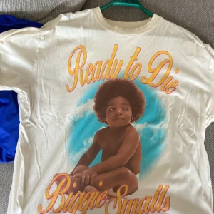 Vit t-shirt med tryck från The Notorious B.I.G. - Säljer en vit t-shirt med ett ikoniskt tryck av ett barn och texten 'Ready to Die' och 'Biggie Smalls'. T-shirten har en loose fit och är perfekt för fans av klassisk hiphop. Tillverkad av bomull för en bekväm känsla.