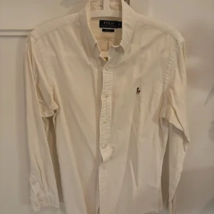 Vit skjorta från Ralph Lauren - Klassisk vit skjorta från Ralph Lauren med broderad logga på bröstet. Skjortan har långa ärmar och knappar framtill. Perfekt för en stilren look.