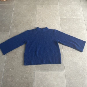 Blå stickad tröja från Indiska - Säljer en snygg blå stickad tröja från Indiska. Tröjan har en enkel design med långa ärmar och en hög krage. Perfekt för kyligare dagar. Passar bra till både jeans och kjol.