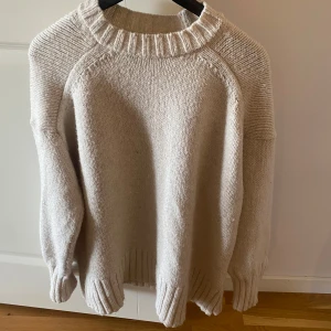 Beige stickad tröja - Mysig beige stickad tröja med ribbade detaljer vid hals, ärmslut och nederkant. Perfekt för kyligare dagar med sin avslappnade passform och långa ärmar.