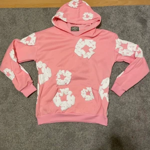 Rosa hoodie från Denim Tears - Säljer en cool rosa hoodie från Denim Tears med unikt vitt mönster över hela tröjan. Den har en bekväm passform med långa ärmar och en praktisk huva. Perfekt för en avslappnad stil!