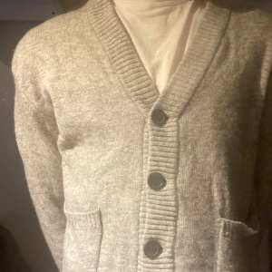 Grå kofta från Lindbergh - Säljer en stilren grå kofta från Lindbergh i lammull. Koftan har en v-ringad design med knappar framtill och ribbade muddar. Perfekt för våren som kommer, vid frågor är det bara att skriva.