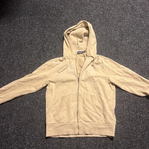 Beige hoodie från Polo Ralph Lauren - Säljer en stilren beige hoodie från Polo Ralph Lauren med dragkedja och klassisk logga på bröstet. Tröjan har långa ärmar och en praktisk huva med dragsko. Perfekt för en avslappnad look.
