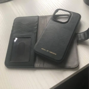 Svart plånboksväska från Ideal of Sweden - Snygg svart plånboksväska till IPhone 15 PRO från Ideal of Sweden. Den har flera kortfack och en avtagbar mobilhållare. Perfekt för att hålla ordning på dina kort och mobilen. Stilren design med magnetlås.