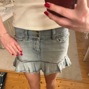 Jeanskjol med volang - Snygg jeanskjol i ljusblått med slitna detaljer och volangkant. Kjolen har två knappar fram och klassiska femficksdesign. Under vänstra fickan nertill är ett hål. Midjemått raktöver: 35cm. Längd: 32 cm. Använd gärna köp nu.💕