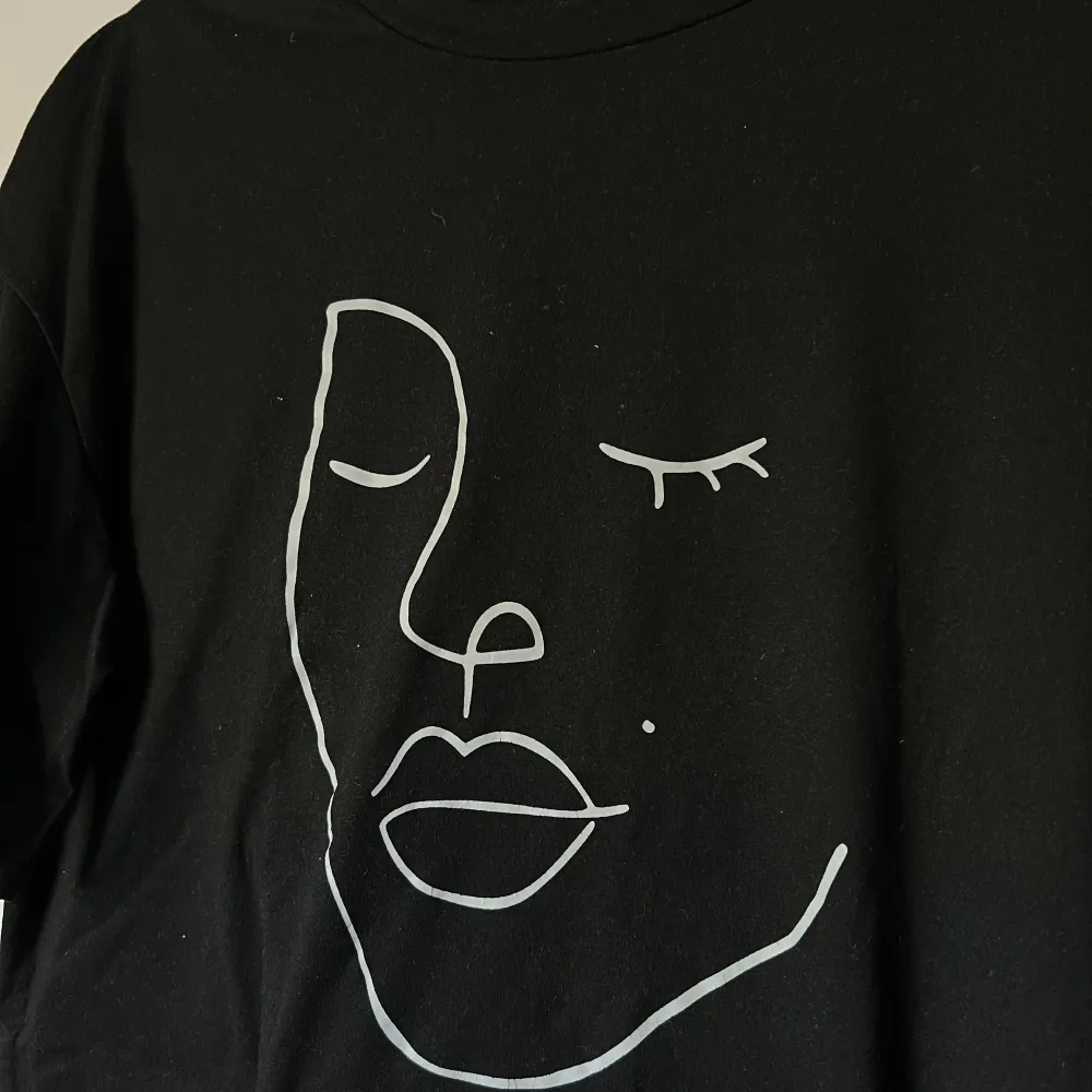 Snygg svart t-shirt med ett stilrent vitt ansiktsmotiv på framsidan. Perfekt för en avslappnad look. T-shirten har korta ärmar och en normal passform. Oversized. Önskar du veta exakta mått? Tveka inte att höra av dig!. T-paidat.