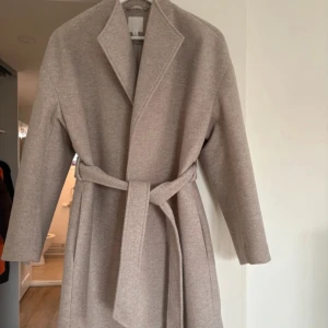 Beige kappa med bälte - Elegant beige kort kappa med bälte och långa ärmar. Perfekt för kyligare dagar med sin stilrena design och mjuka material. Ett måste för garderoben! Är i storlek XS men passar XS-M