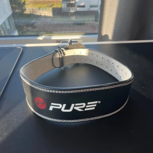 Svart träningsbälte från Pure - Säljer ett svart träningsbälte från Pure med vit insida och dubbla spännen för extra stöd. Perfekt för styrketräning och tyngdlyftning. Bältet har en robust design med vita sömmar som ger en snygg kontrast. Passar bra för dig som vill ha extra stabilitet under träningen.